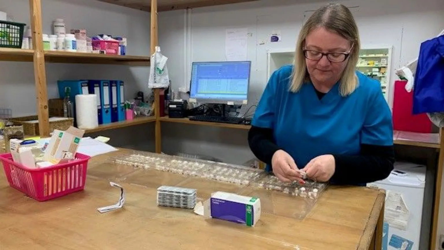A woman with shoulder length blonde hair, who is wearing dark rimmed glasses, is looking down on a table as she sort tablets into different compartments in blister packs. She is wearing a blue coloured tunic over a black top.
