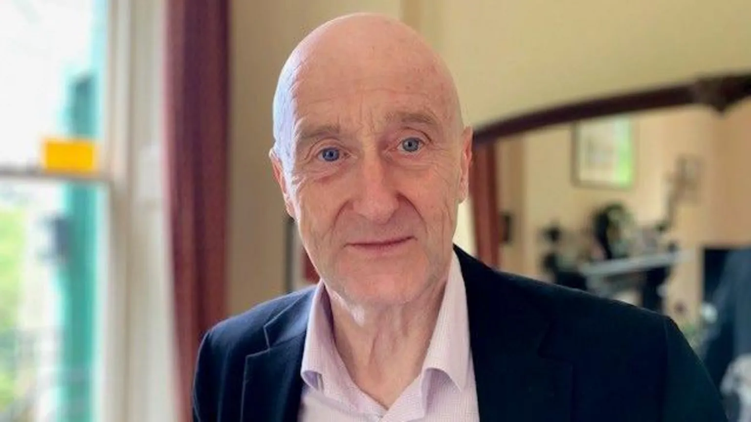 A man is looking at the camera. He is wearing a pale pink shirt and a navy blazer and is standing in front of a window and a mirror.