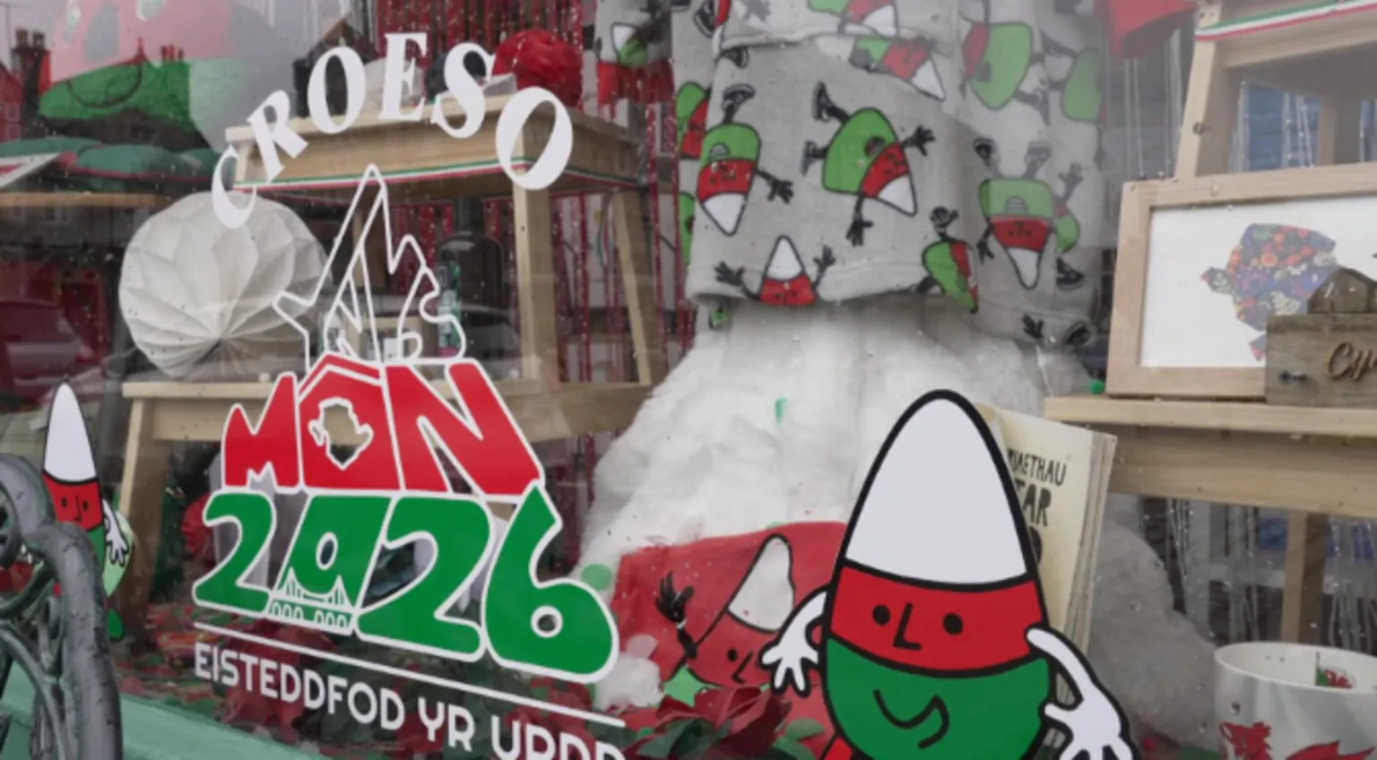 A shop window decorated with the festival's logo which is white, green and red. It has the name of the festival, the year 2026 and the word croeso, plus a little cartoon style character to the right in green, red and white stripes.