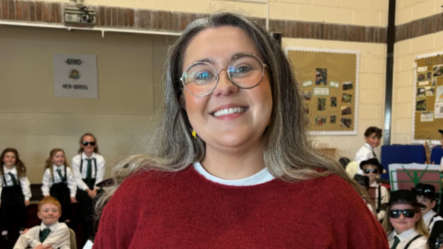 Bronwen is a young woman with dark hair past her shoulders and large round, wire-rimmed glasses. She is wearing a red round-neck sweater with a white t-shirt underneath. She is smiling at the camera.