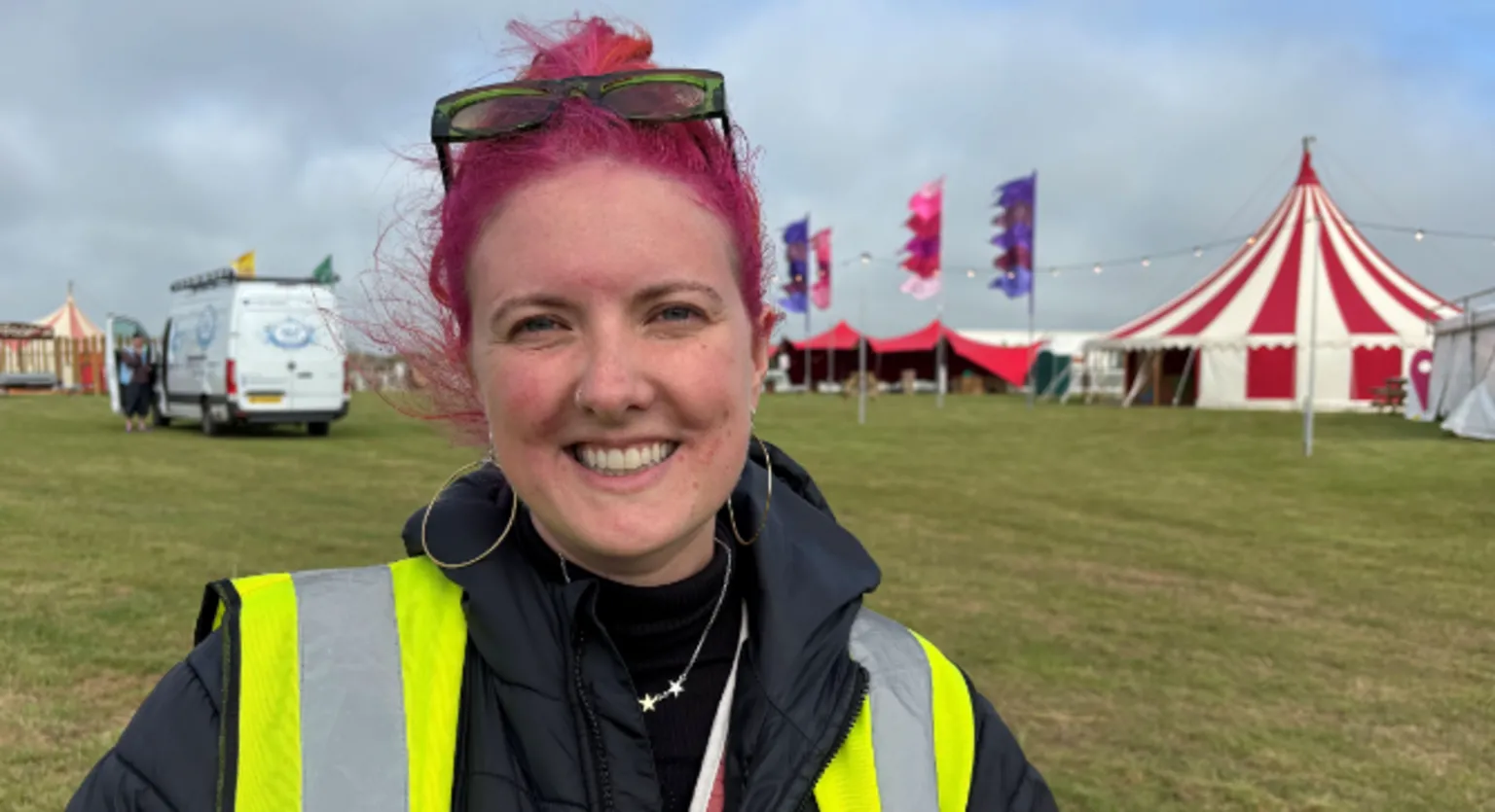 Llio is standing at the festival site; behind her is a red and white striped tent, festoon lights and pink and purple flags. She has bright pink hair and green sunglasses on top of her head. She has large silver hoops in her ears and is wearing a silver necklace featuring stars, a yellow high-vis vest and a blue coat.