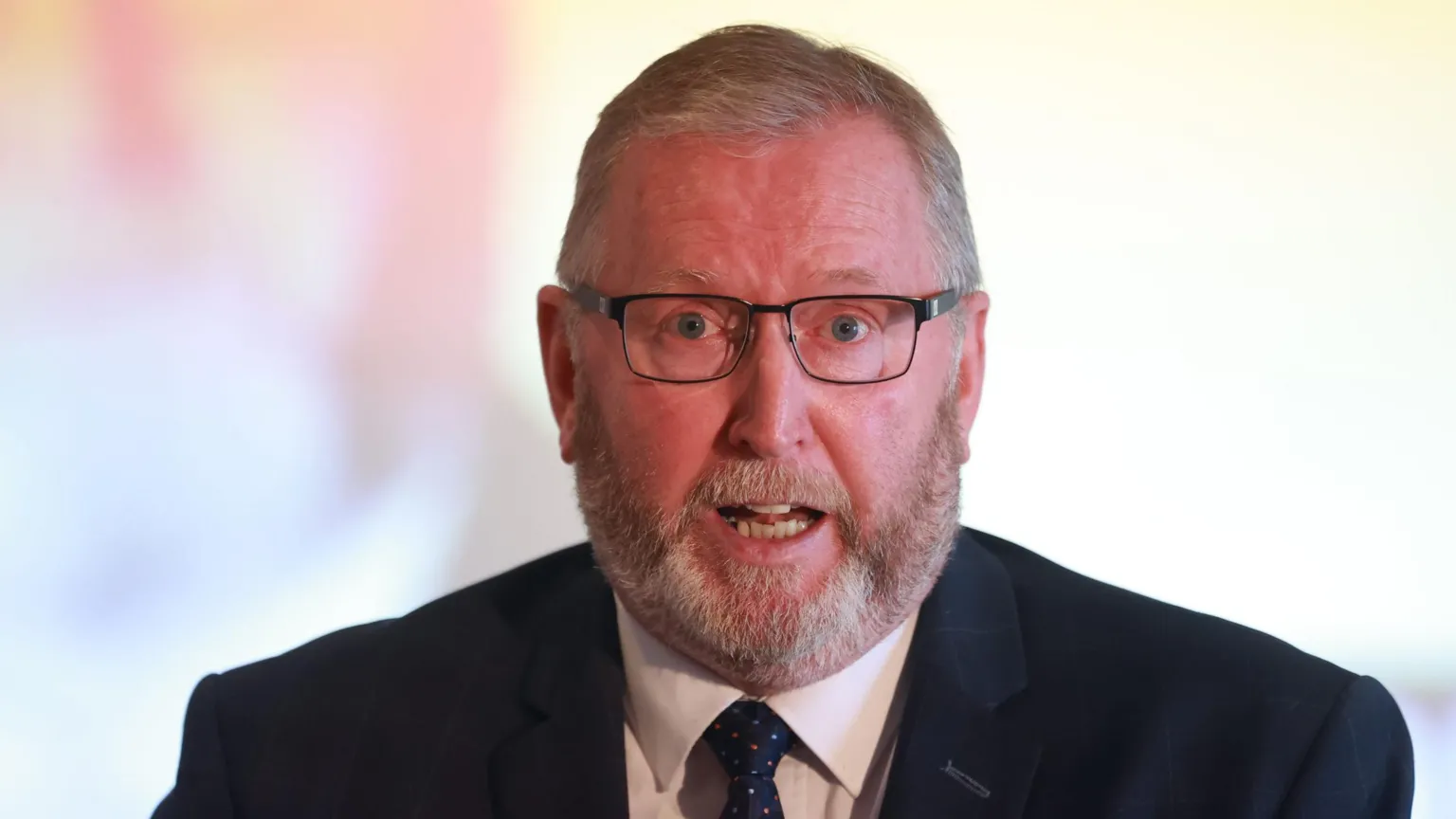Doug Beattie has grey hair and a grey beard. He is wearing wired framed, rectangular frames, a navy jacket, white shirt and navy tie.