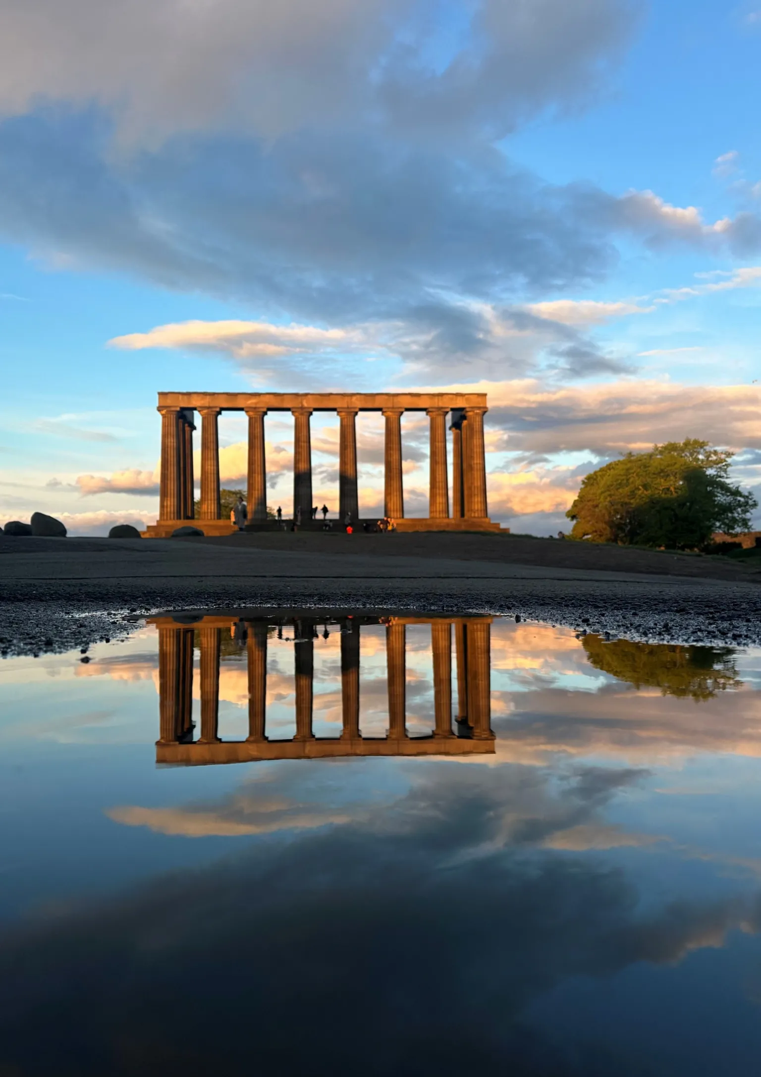 Leora Soibelman Monument with tall columns silhouetted against a partly cloudy sky at sunset, reflected in a puddle in the foreground.