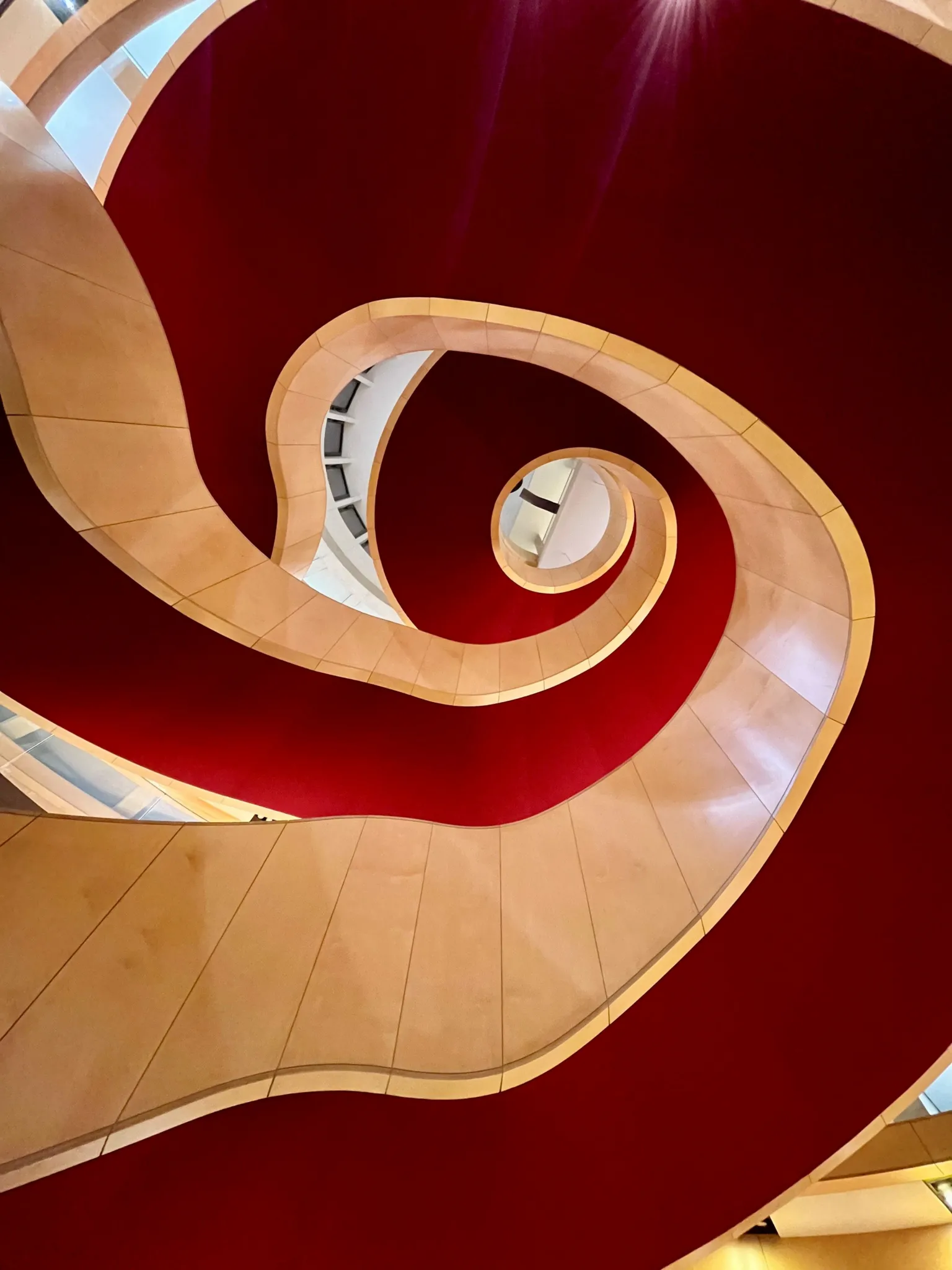 Susan Morrison Looking up through a curved spiral staircase with smooth, cream-colored walls set against a bold red backdrop, forming a flowing, abstract pattern