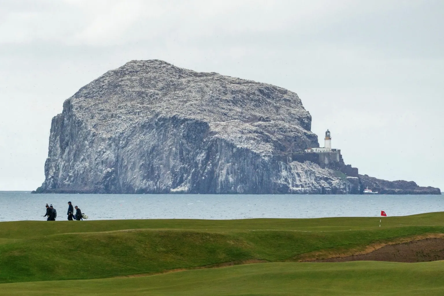 Chris Bell Large rocky island covered in seabirds rising from the sea, with a lighthouse perched on one side. In the foreground, a grassy golf course with a flag and several people walking along the links.