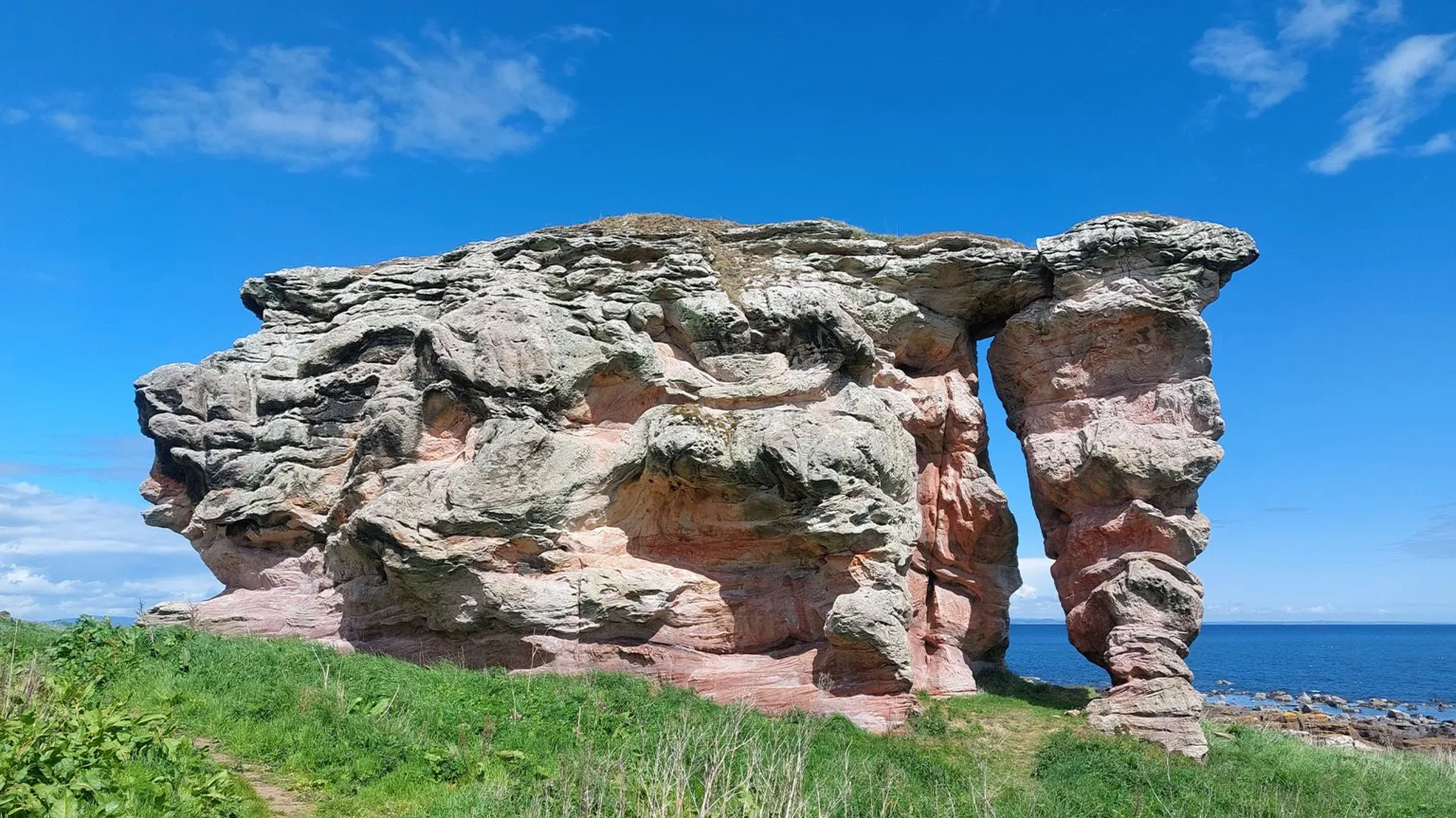 Ian Marshall Large weathered rock formation with layered red and grey tones, shaped into a natural arch on a grassy coastal cliff, with the sea visible beyond under a clear blue sky.