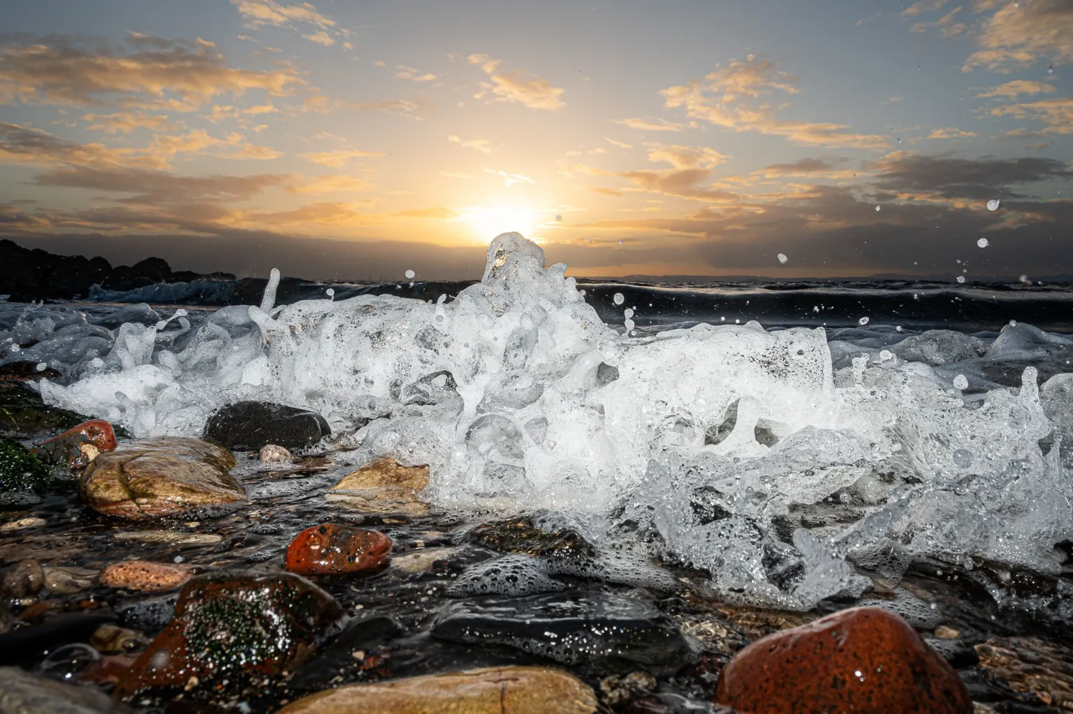 Stephanie Virot Close-up of waves breaking over smooth shoreline stones at sunset, with water splashing into the air and the sun low on the horizon casting warm light through scattered clouds.