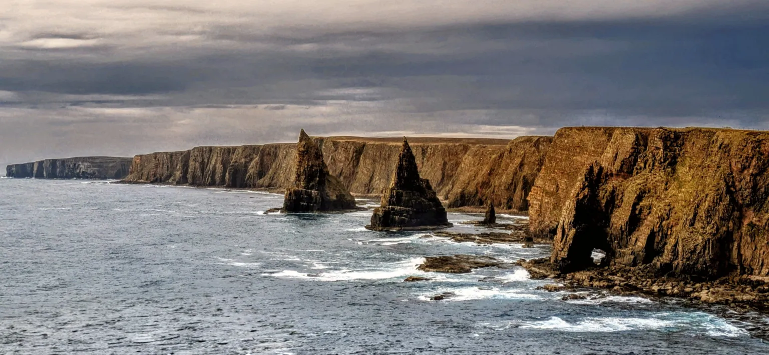Gareth Talbot Dramatic coastal cliffs with two tall sea stacks rising from the ocean. Waves break against rocky shores under a cloudy sky, with a natural arch visible in the cliff face.
