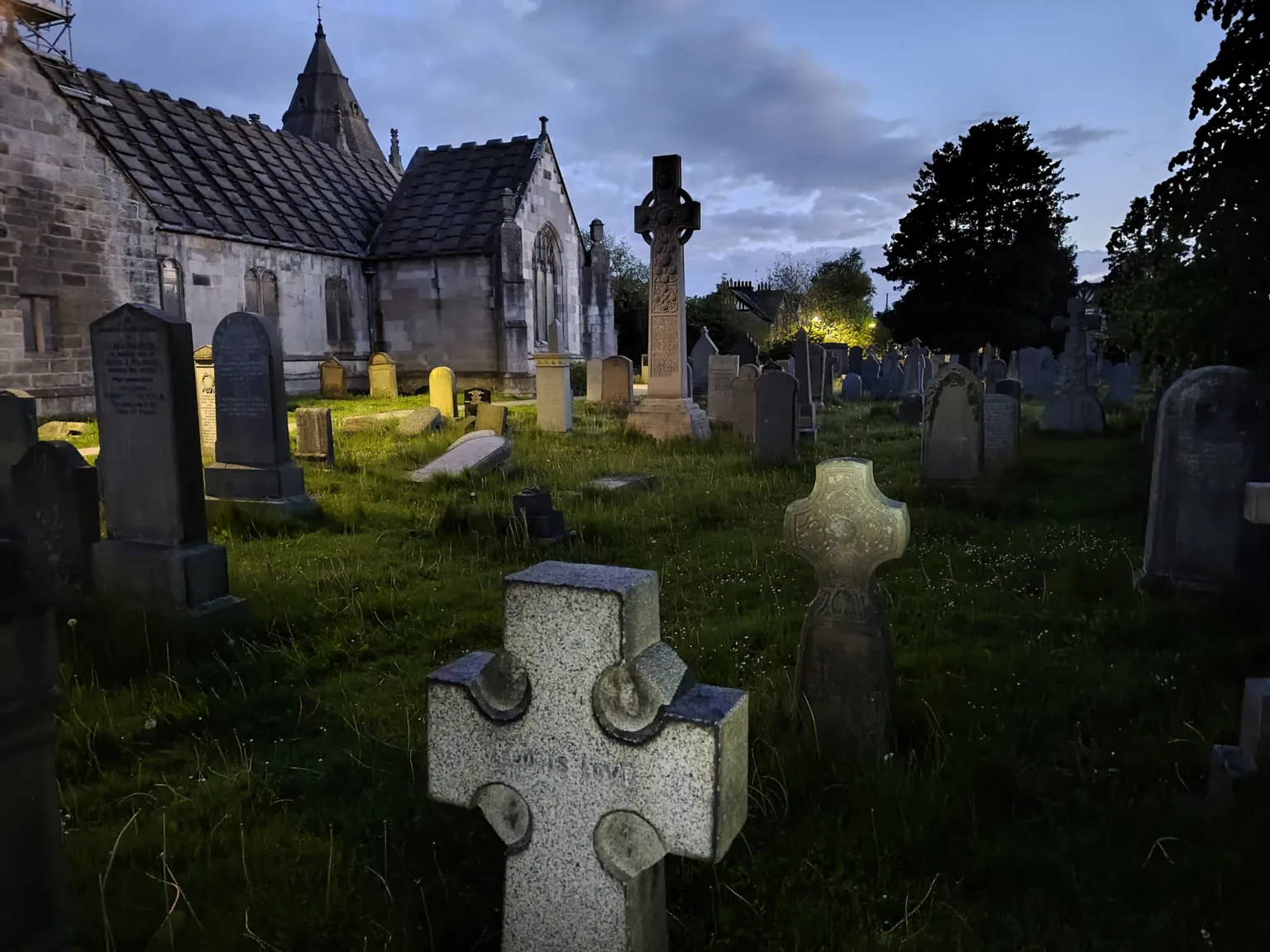 Kevin O'Kane Graveyard at dusk with rows of headstones and a stone church in the background. A tall Celtic cross monument stands among the graves, with soft lighting from nearby buildings under a cloudy evening sky