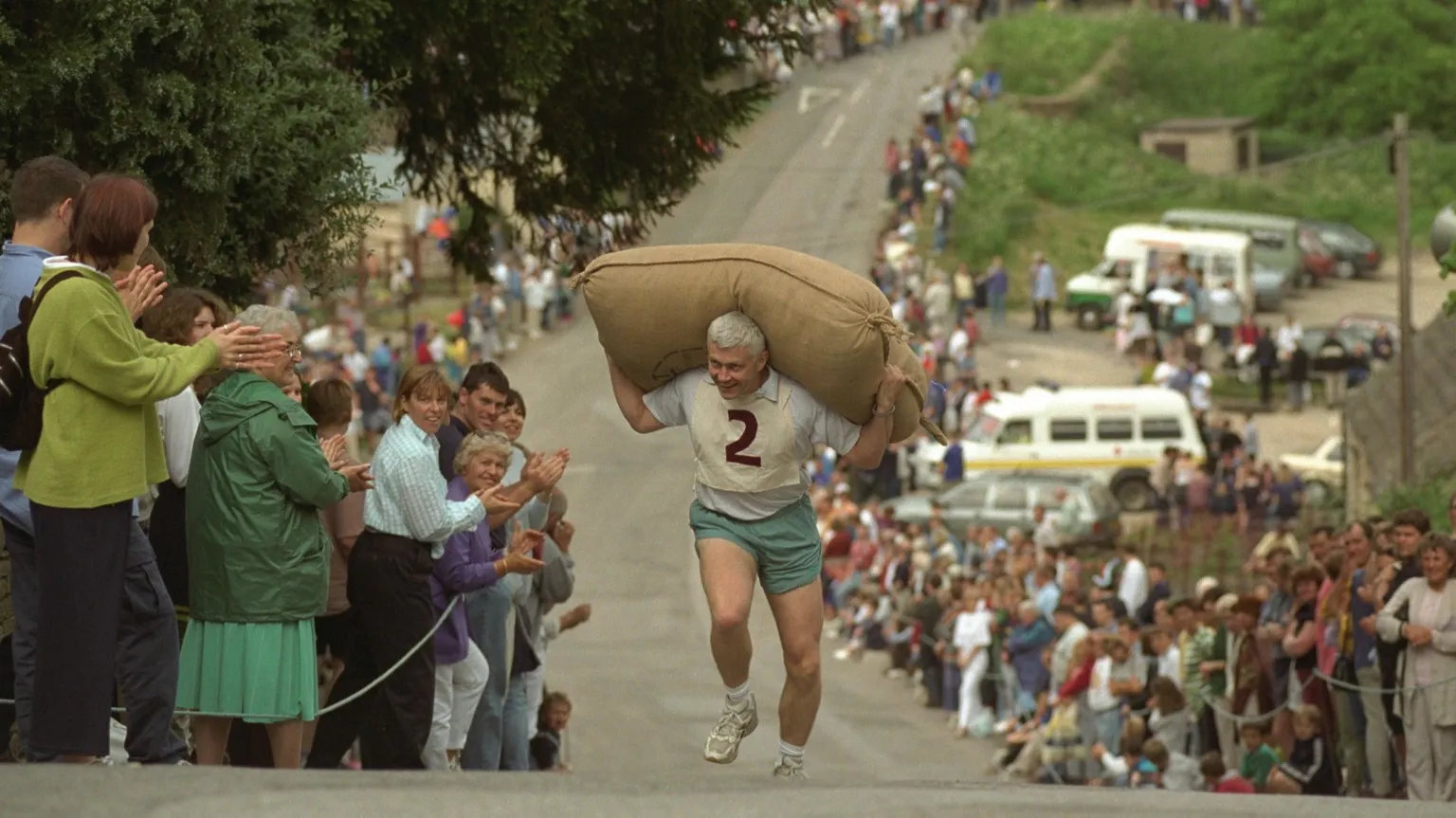  A man with grey hair and a bib with the number two is running up a hill with a large brown sack on his shoulders. The street is lined with crowds of people.