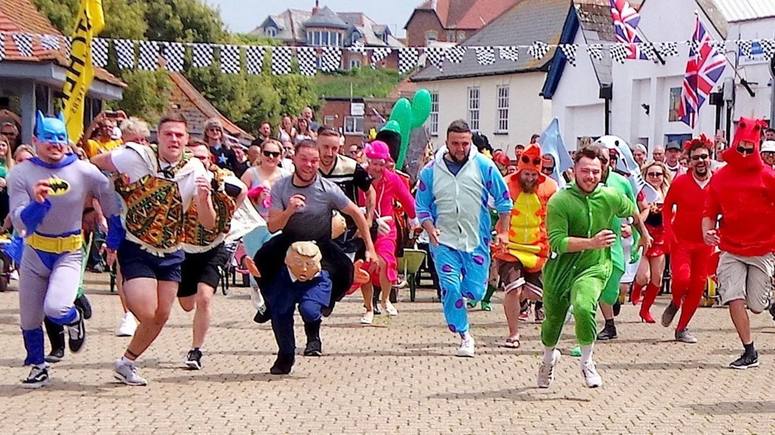 Watchet wheelbarrow races A line of competitors in the Watchet wheelbarrow races smile and shout as they sprint towards the camera. Many of them are in fancy dress