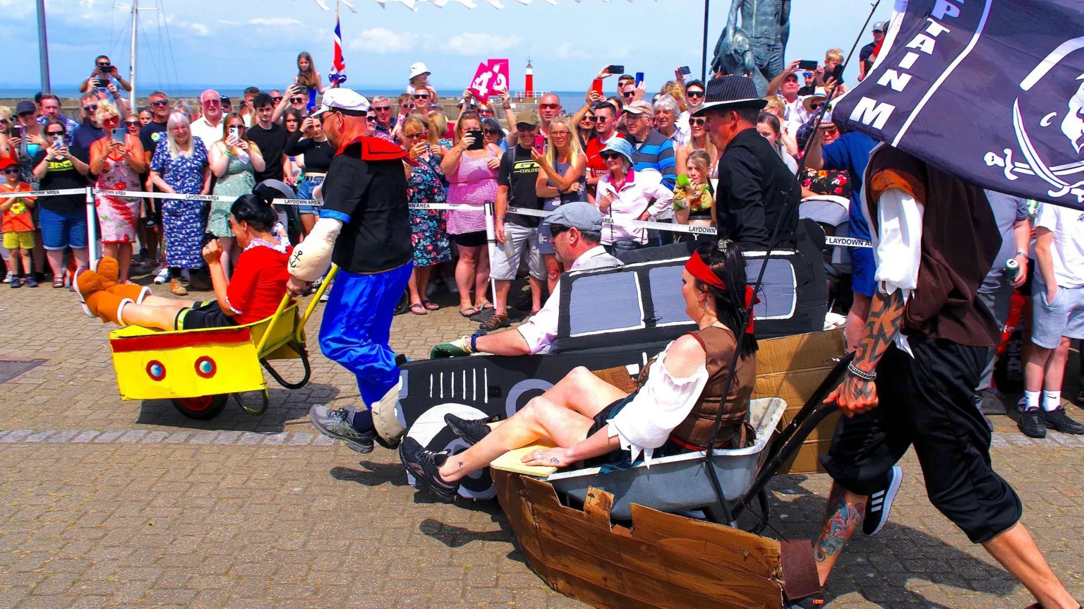 Watchet Wheelbarrow Race Three different sets of competitors take part in the Watchet wheelbarrow races, with the people in the wheelbarrows wearing different types of fancy dress. It is a sunny day, and dozens of people are watching from the roadside
