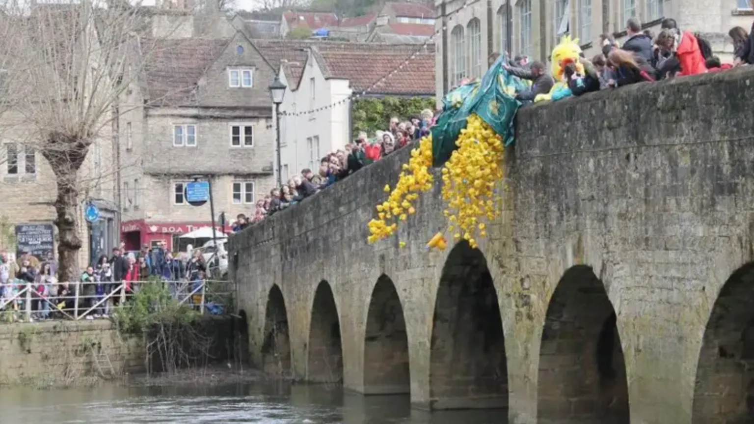 Bradford-on-Avon Town Council Crowds of people stand on an old bridge in Bradford-on-Avon as bags of rubber ducks are thrown into the water. Near the bridge, people stand and watch.