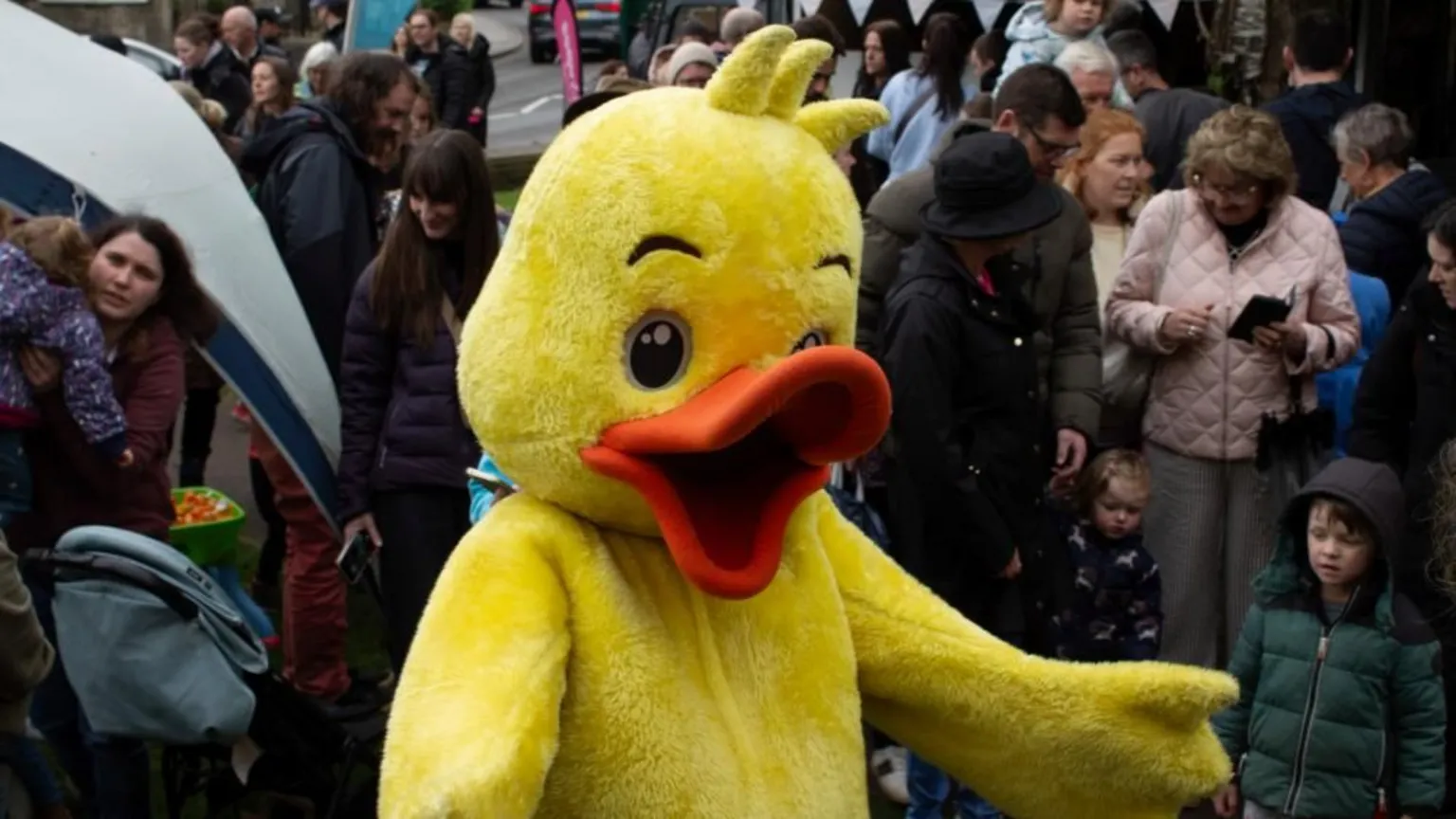 Bradford-on-Avon Town Council Someone wearing a rubber duck suit with a yellow body and orange beak. They are stood with their arms out in a crowd.