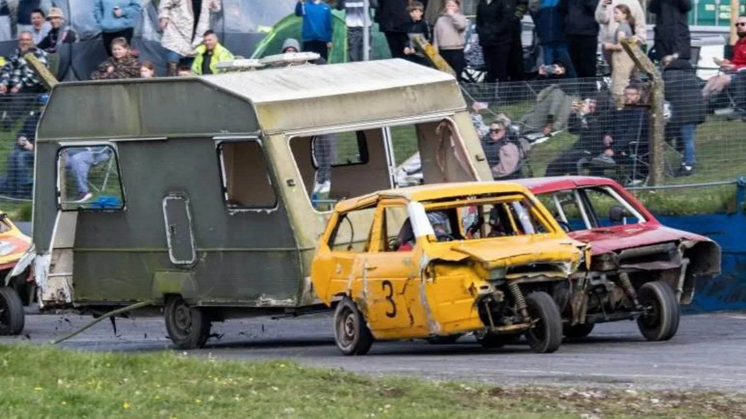 Lime Jelly Photography Beat up three-wheeled cars and a green caravan are driving around a track with a small crowd behind on a grassy bank.