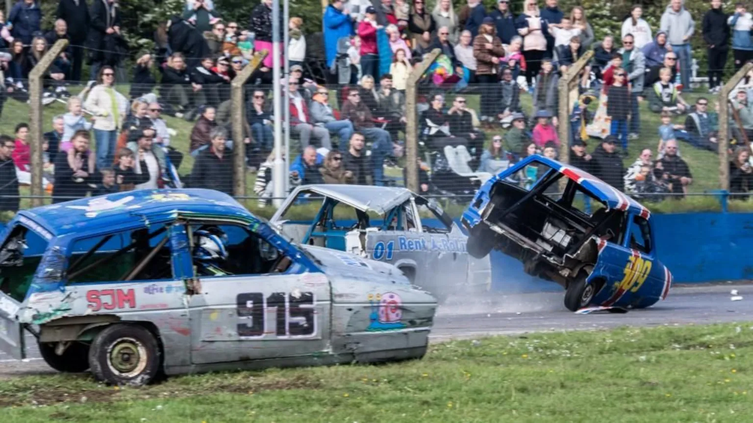 Lime Jelly Photography Beat up cars drive round a track with the crowd behind a fence in the background. One of the cars is driving onto the grass while another has two wheels up in the air.