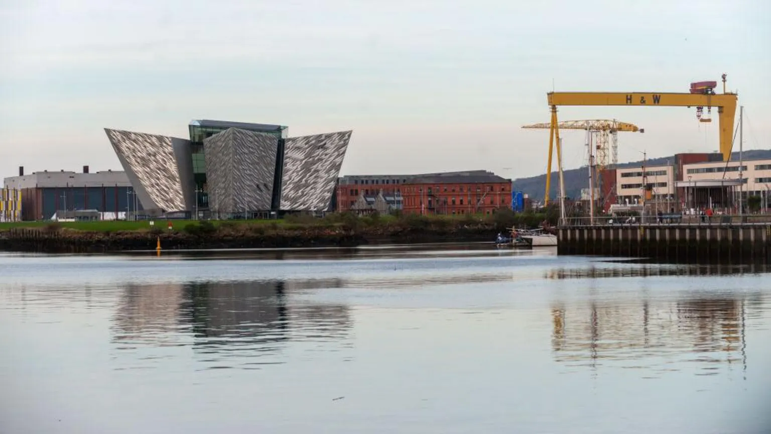 The massive silver Titanic Experience building in Belfast. It has many points and is against a blue sky, surrounded by cranes and water.