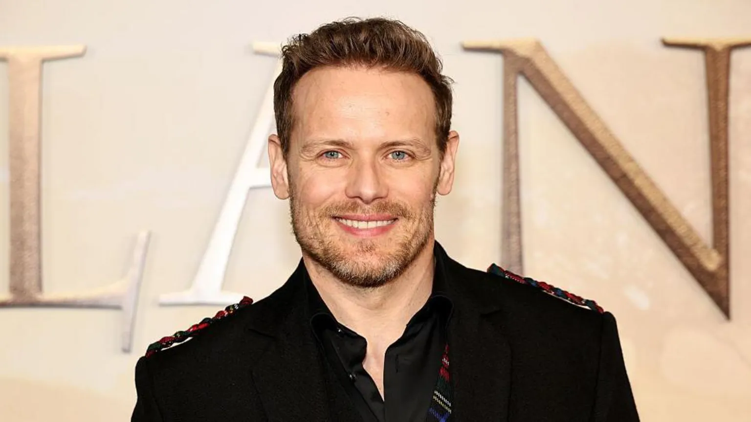 Sam Heughan with short brown hair wearing a black jacket, shirt and red and green tartan waistcoat smiling at the camera.