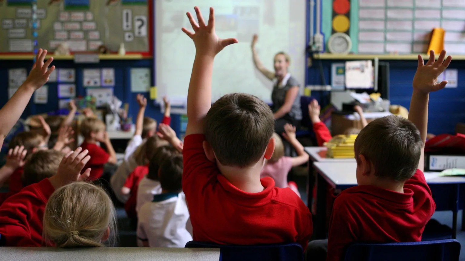 The back of children's heads sat in a classroom. They wear red polo shirts and many of them have their hands raised. A woman with blonde hair is sitting at the front of the classroom facing the pupils.