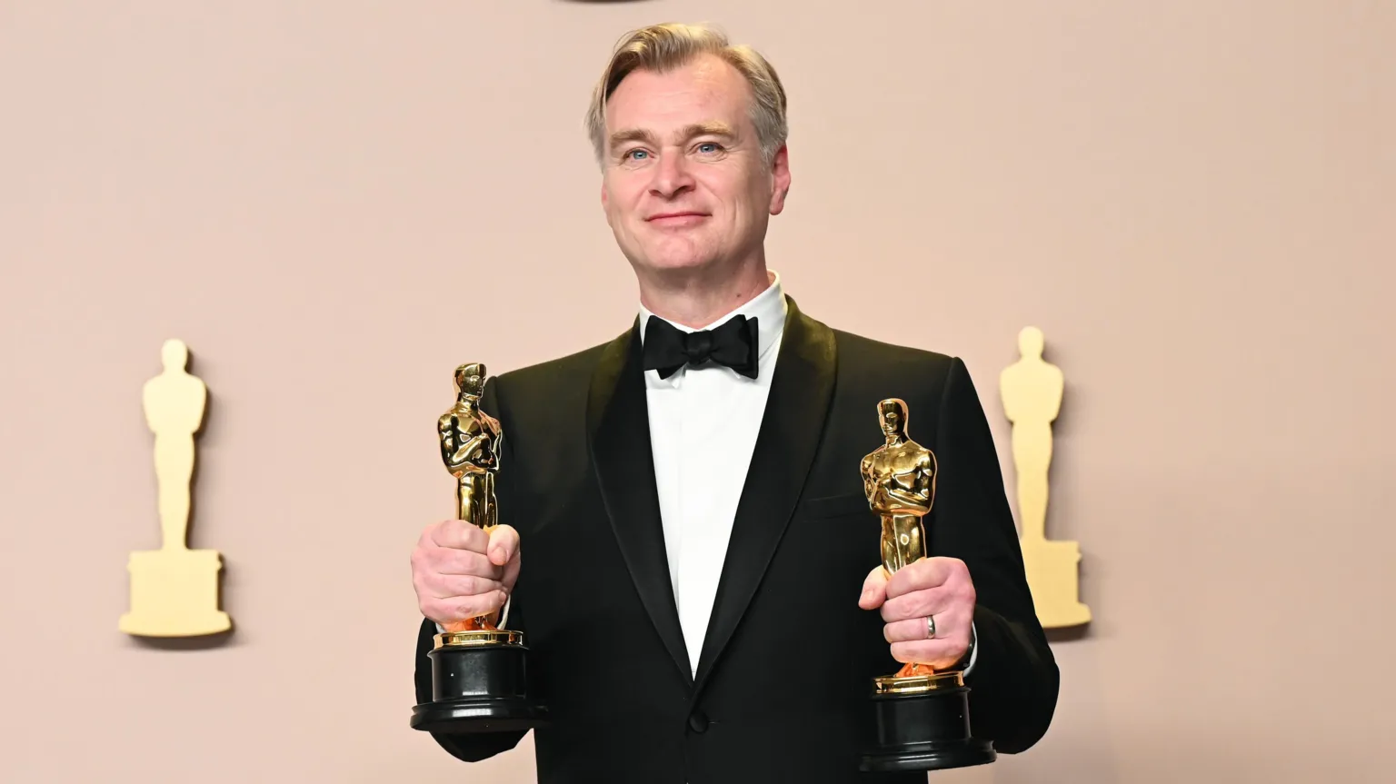  Christopher Nolan smiling in a black bow tie and dinner jacket, holding an Oscar statuette in each hand, in front of an Oscars backdrop