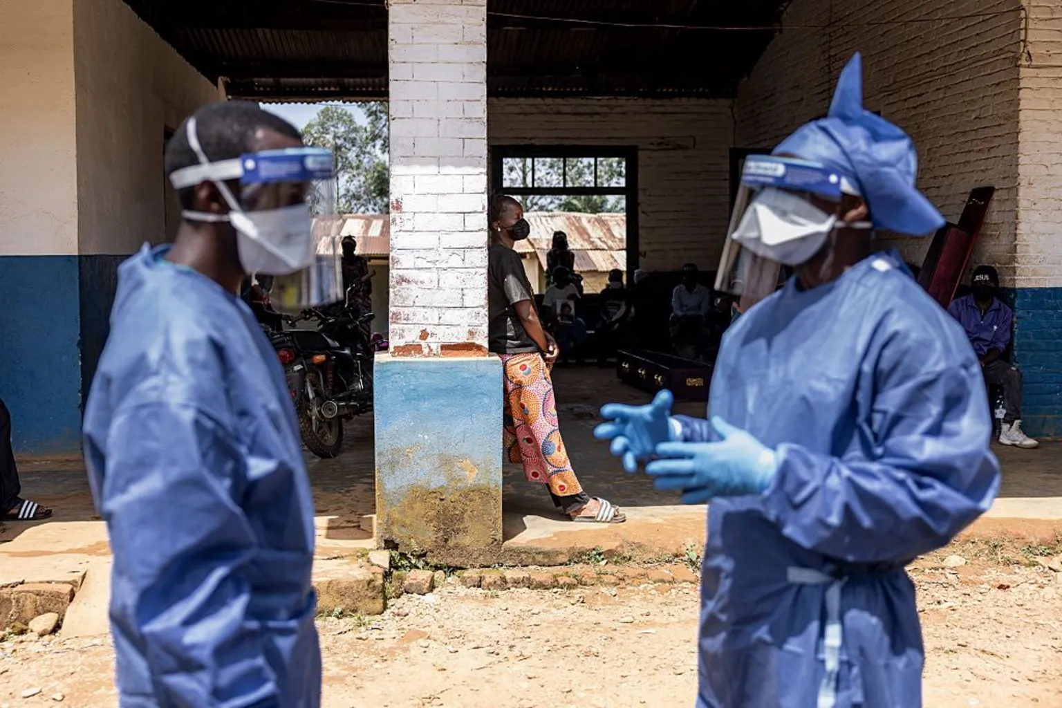  The image shows a partially open building with a shaded interior and a dirt ground outside. In the foreground, two individuals are standing and wearing full protective gear, including blue gowns, gloves, face masks, and clear face shields. One individual appears to be gesturing with gloved hands, while the other faces toward them, suggesting a conversation or coordination. In the background, several people are sitting or standing under the roofed area, some near parked motorcycles and a wall with a window. 