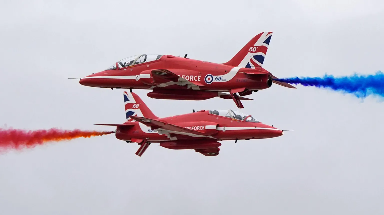  Two red Royal Air Force planes fly through the sky. The plane on the bottom shoots out red smoke to the left, while the plane on the top shoots out blue smoke to the right.