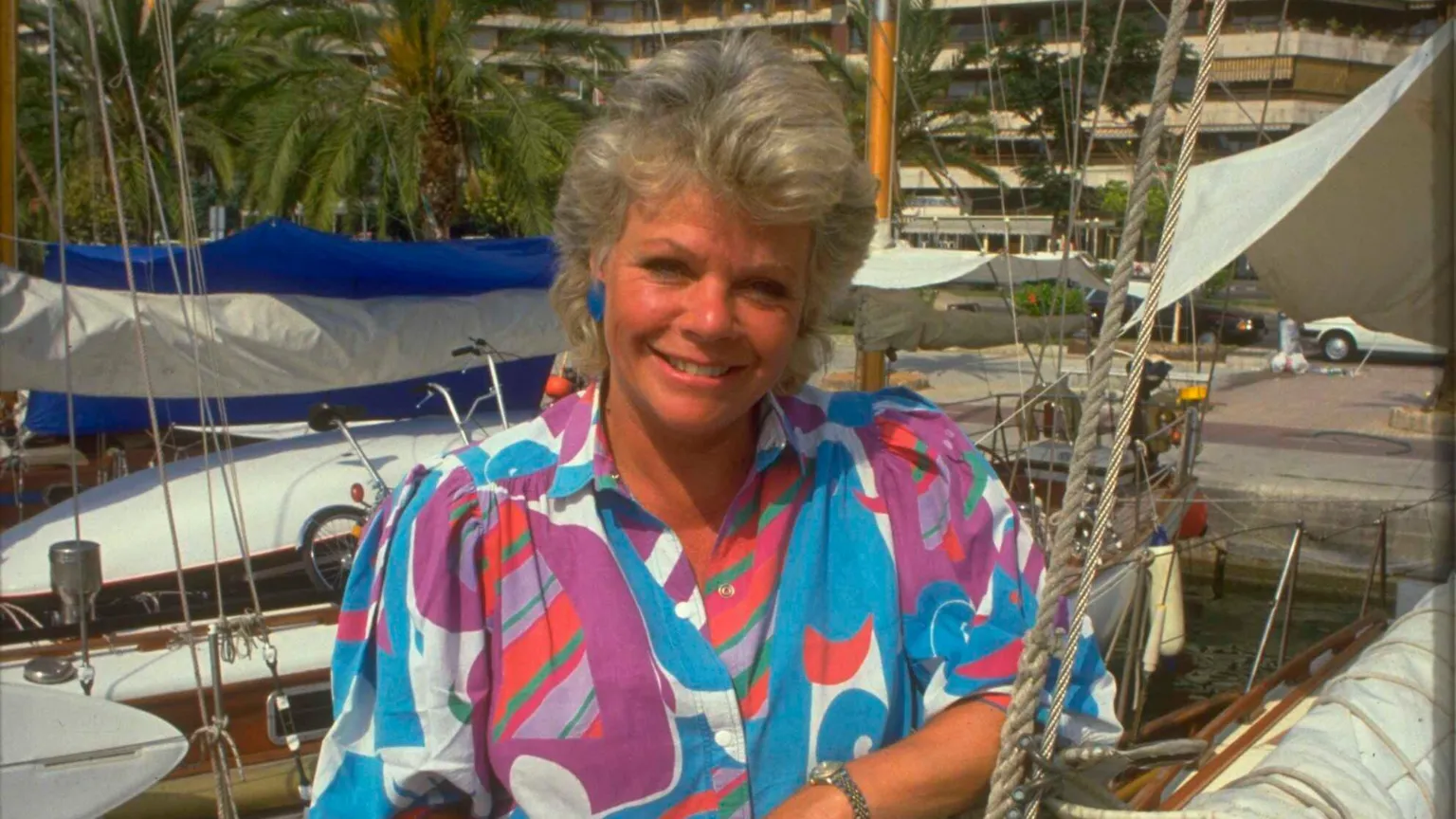 Judith Chalmers in a bright multi-coloured blouse in front of a Mediterranean harbour on Wish You Were Here?