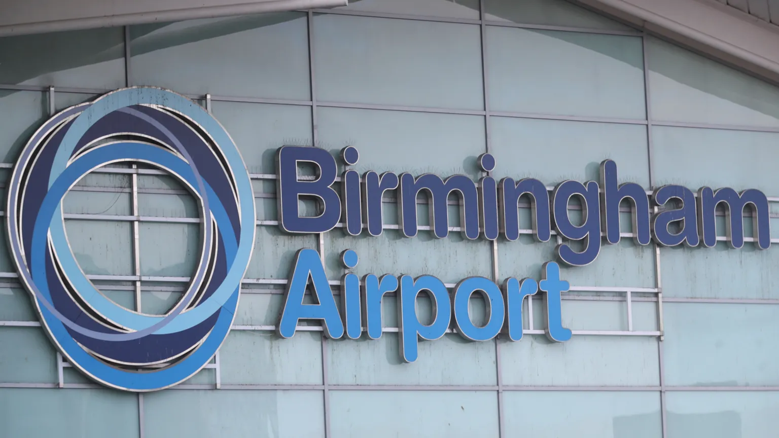  Birmingham Airport is written on the side of a building with a fogged glass exterior. The words are in dark blue and light blue lettering and there is a circular symbol to the left of the words. 