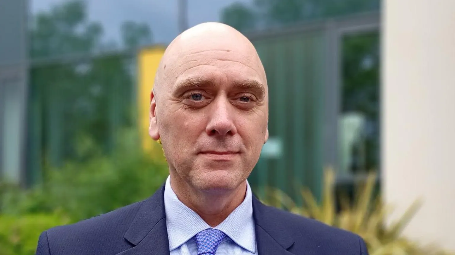 Trevor has a wet shaven bald head. He is wearing a navy suit with a pale shirt and blue tie. 