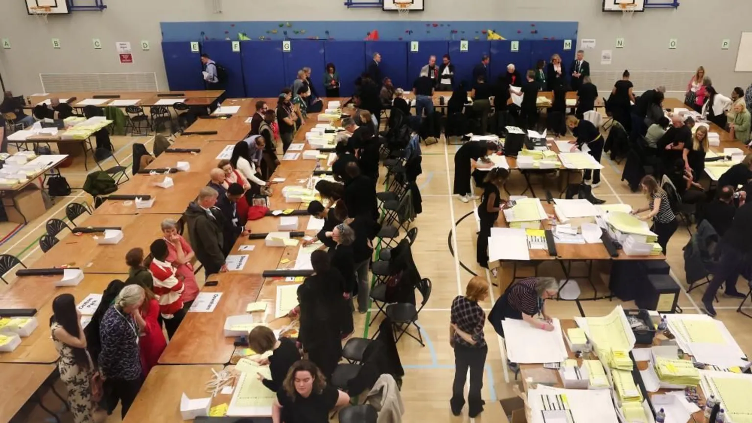 EPA/Shutterstock Many people sit in rows of desks in front of piles of papers. The room appears to be a gymnasium with playing field markings on a brown wooden floor.
