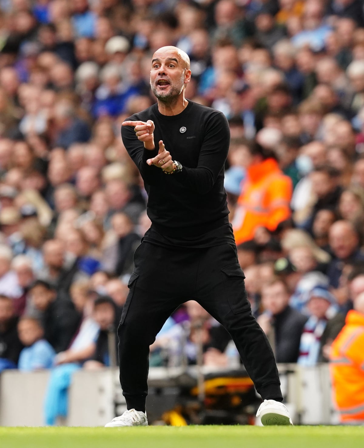 Pep Guardiola on the sideline of the pitch, dressed in black combat-style trousers with white trainers