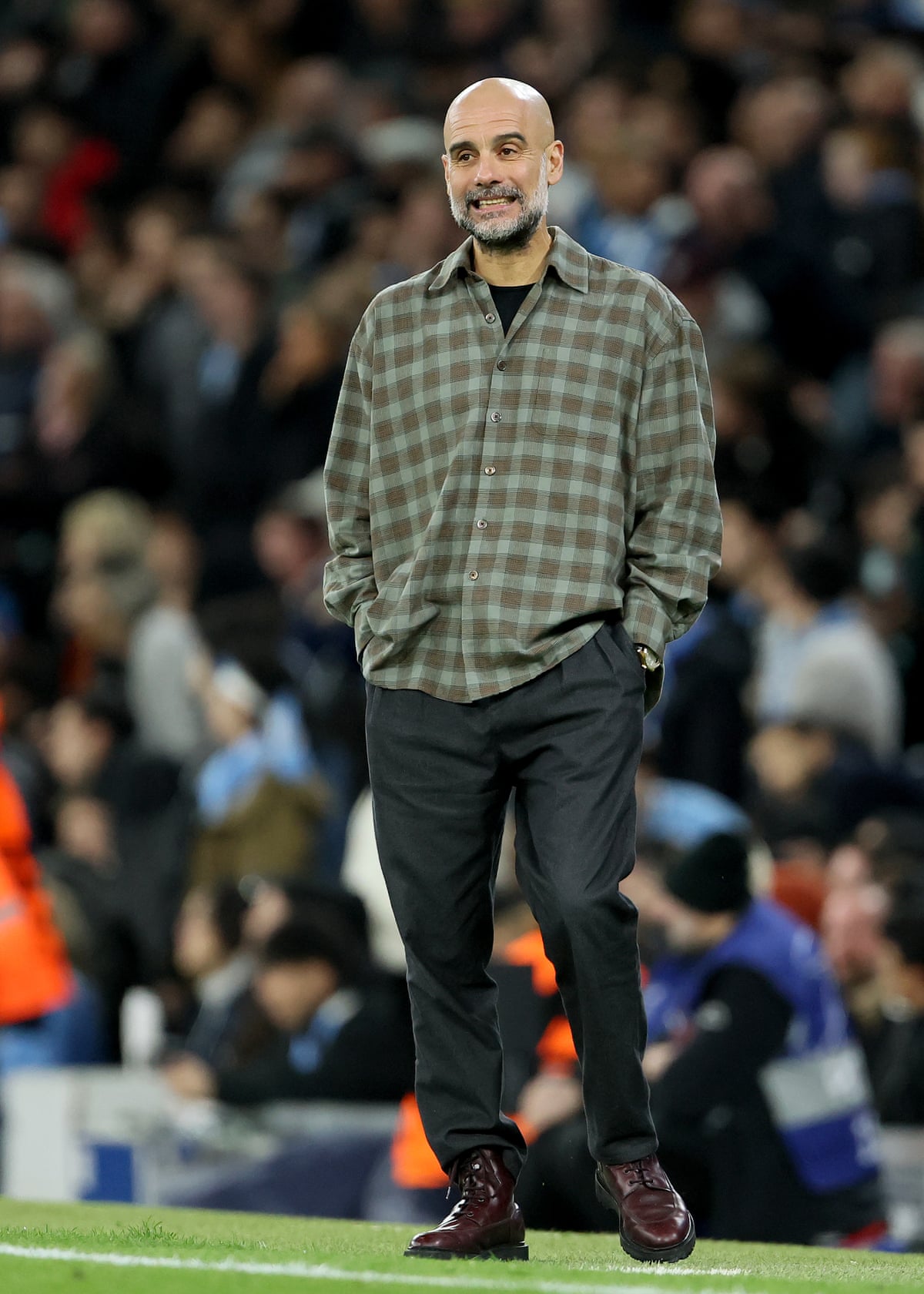 Pep Guardiola wearing a checked shirt and black trousers on the sideline of the pitch