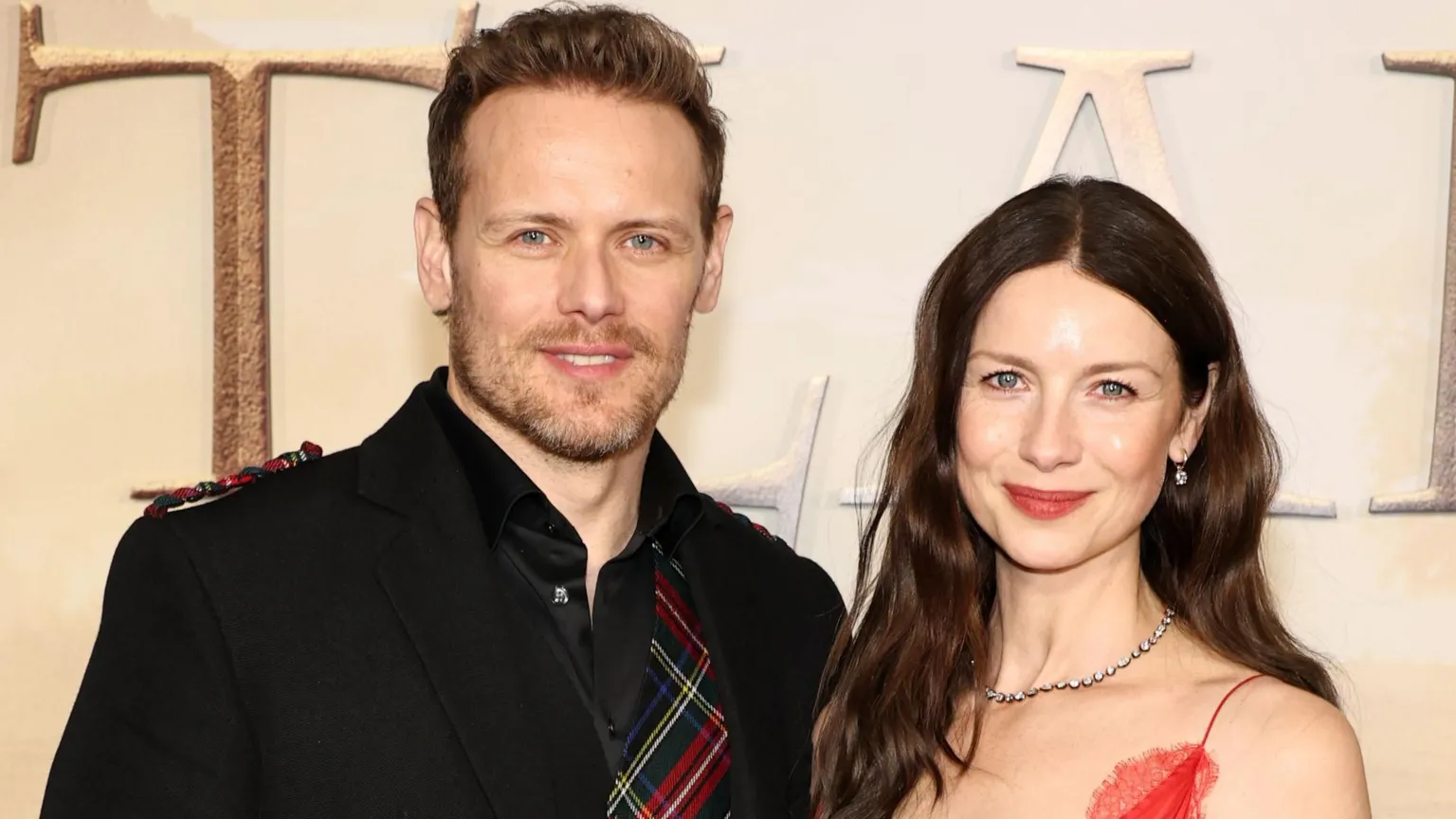 A red carpet premiere - Sam Heughan stands wearing a kilt and tartan patterned shirt, while Caitriona Balfe wears a red dress.