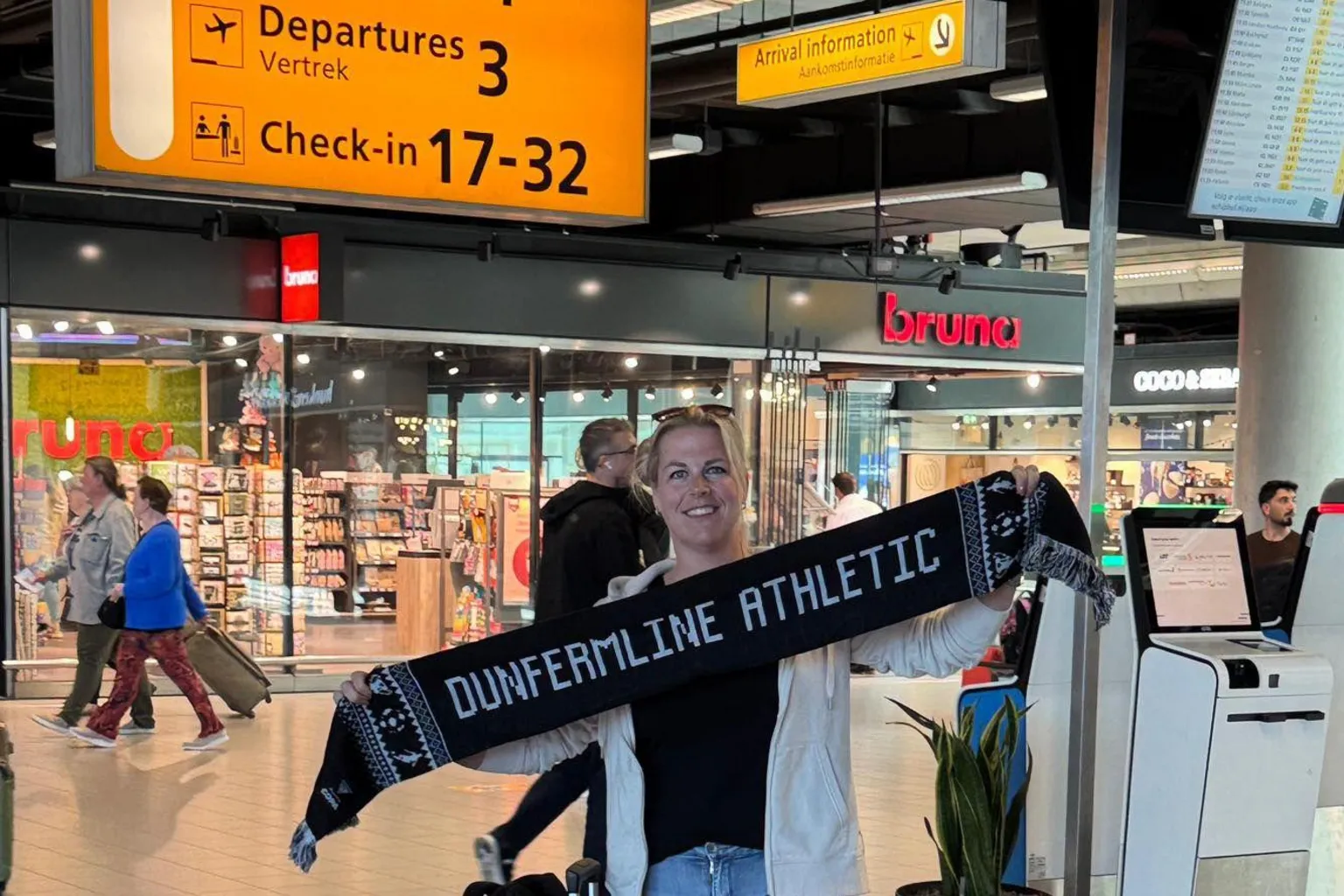 Amanda Thompson A woman with blue jeans stands in an airport with a suitcase holding up a black and white Dunfermline Athletic scarf