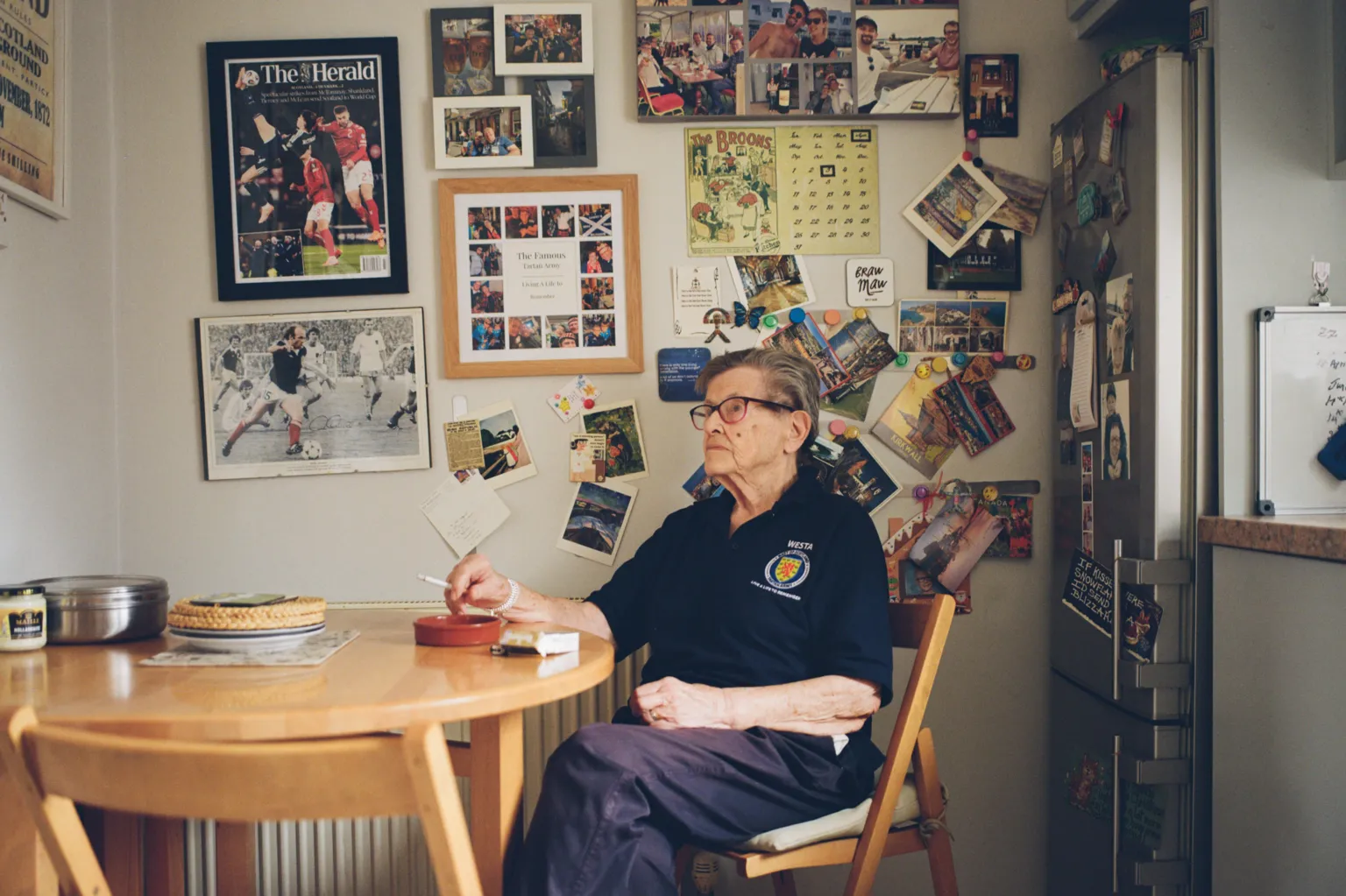 Craig Mcintosh Moira sitting at the table of her kitchen, the wall behind her is covered with framed photographs.