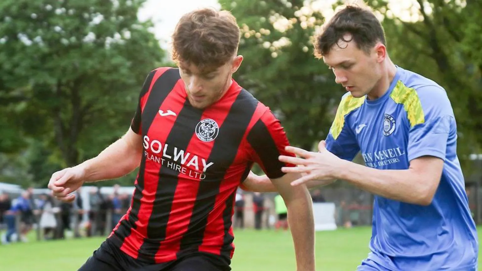 David McDonald A footballer with red hair and a beard with a red and black striped top shields the ball from a man in a blue shirt with gold stripes on it