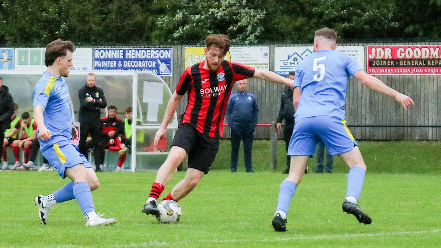 David McDonald A footballer in a red and black striped top and black shorts has the ball at his feet while two players in blue kit with yellow shoulder stripes close in on him