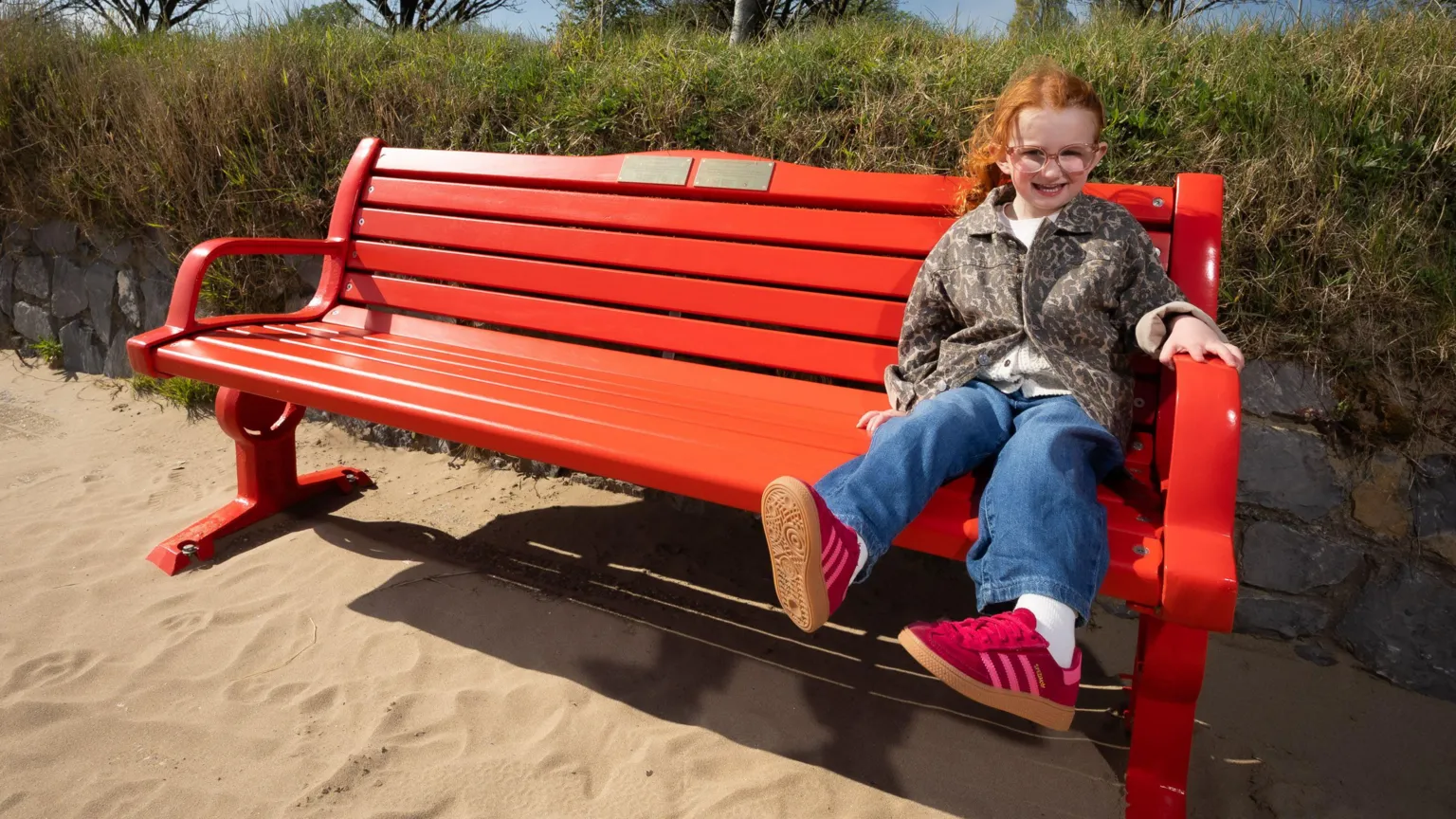 British Heart Foundation A little girl with red hair and glasses wearing a white t shirt and leopard jacket, and pink adidas trainers, sat on a red bench with a gold plaque on it. Behind her is a green bush and in front is a sandy beach. It's a sunny day and she is smiling and looking at the camera.