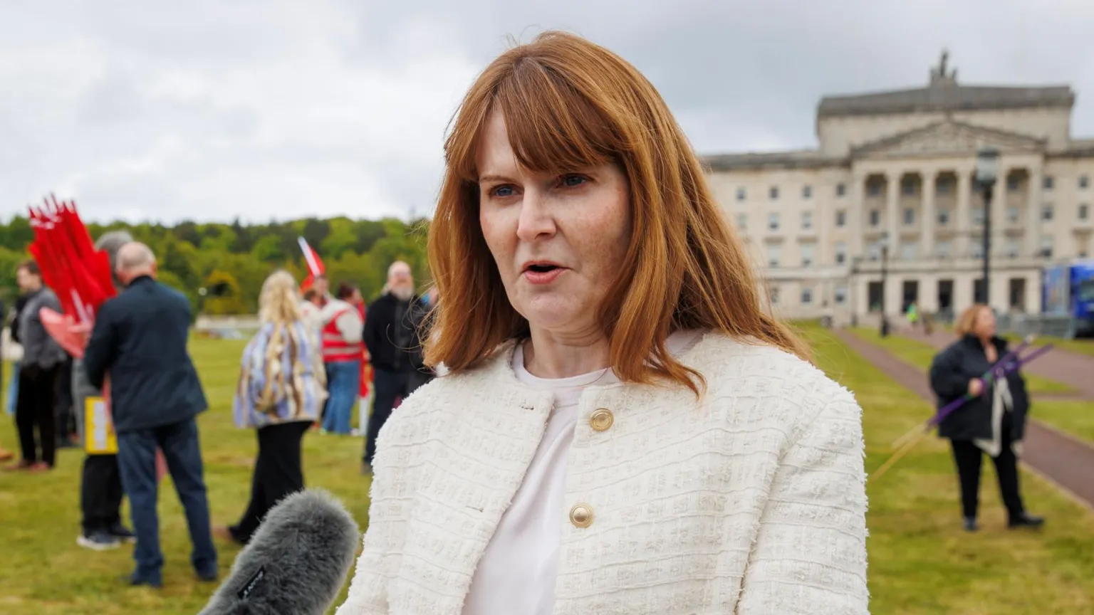 Caoimhe Archibald speaking into a microphone. She is wearing a cream jacket with gold buttons. She has long red hair. She is standing outside Stormont parliament buildings.