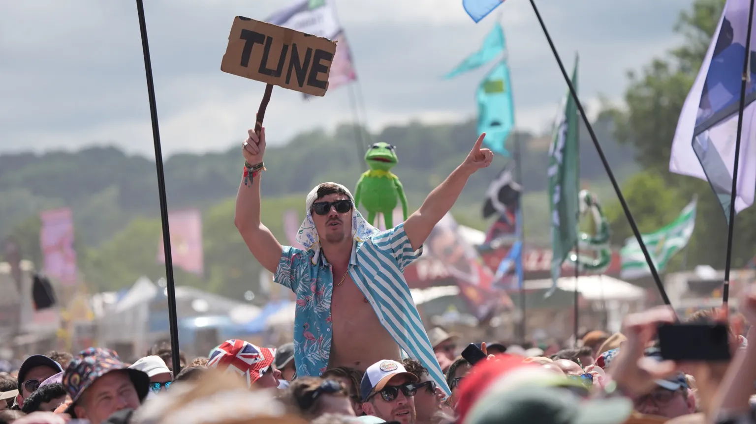 A man in an open blue and white shirt sits on the shoulders of a friend at Glastonbury festival with a sign that says