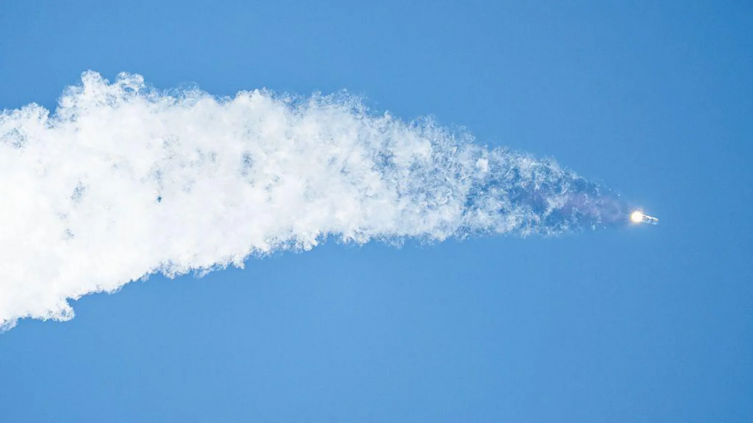  A space rocket flies upwards into a clear blue sky, leaving a huge grey trail behind it in a widening streak.