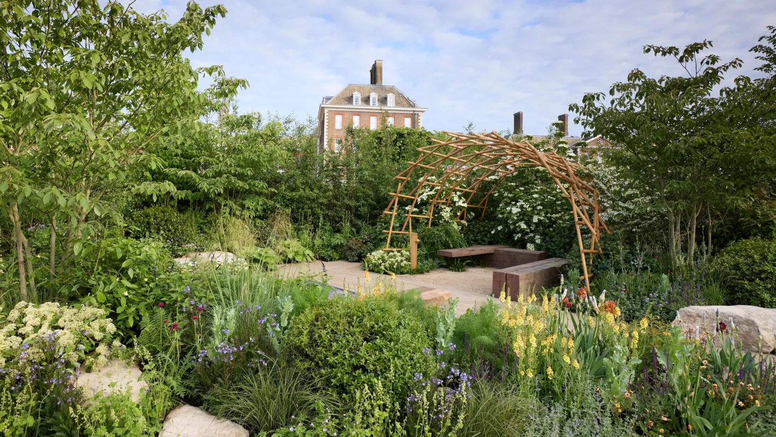 Gary Morrisroe A wide shot of the award-winning Chelsea Flower Show garden, with a partial view of the Royal Hospital Chelsea in the background. The garden has a large seating area, featuring a curved, wooden pergola over three large wooden blocks used as benches. There are green shrubs around the edges of a walkway and yellow, orange, red and purple flowers line the front of the picture.