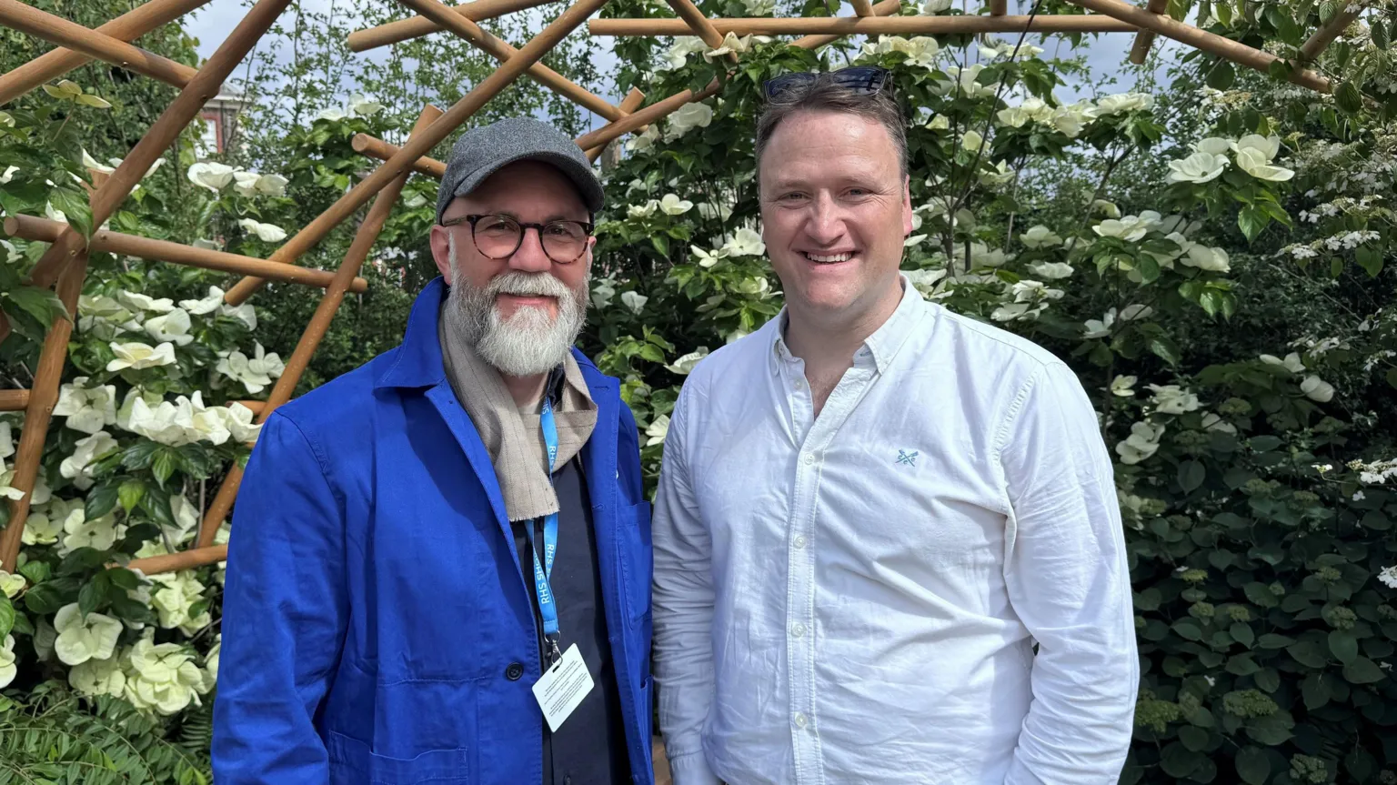Gary Morrisroe Rob Hardy poses for a photo with David Maxwell while standing under a curved wooden pergola, in front of shrubbery with large white flowers. Hardy is wearing a grey baseball cap, dark-rimmed glasses. a blue coat and a navy jumper. David is wearing a white, open-necked shirt.