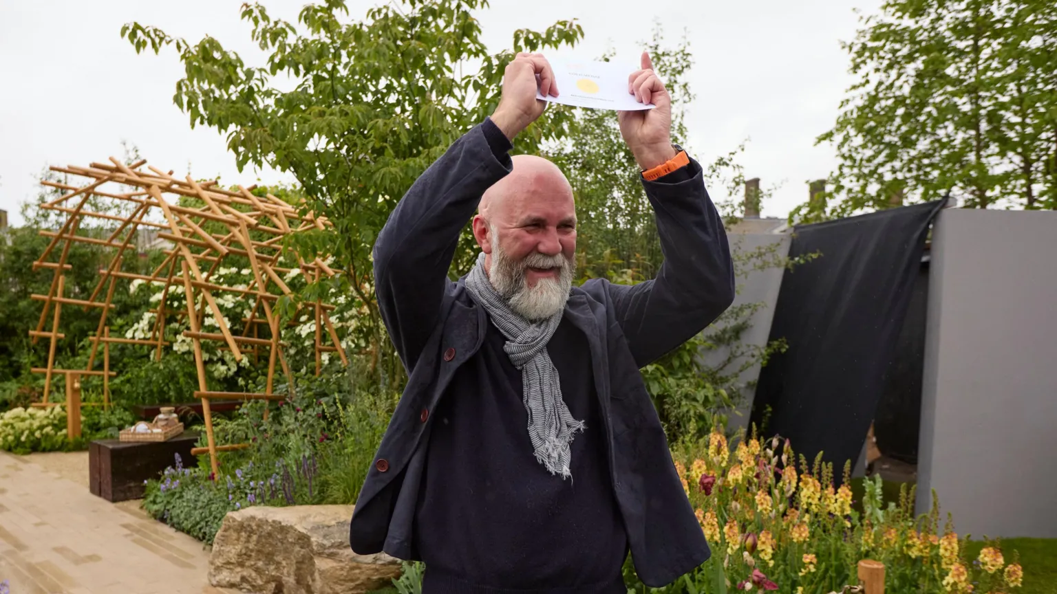 Gary Morrisroe Rob Hardy, a man with a shaved head and white beard, holds a gold medal presentation card above his head while standing in his award-winning garden. He is wearing a navy jacket, a navy jumper and a navy and white scarf