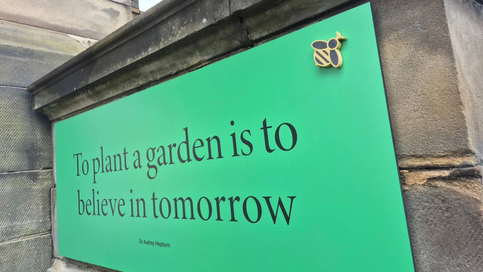 A large sign reading to plant a garden is to believe in tomorrow, with black lettering on a green background. A small model of a bee has been attached to the top right hand corner.