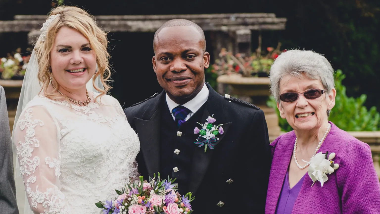 Innovation Photography Charmain, wearing a white wedding dress and carrying a bouquet, and Eric Adusah, wearing a kilt, at their wedding, with Elma Adams on the right of the picture.