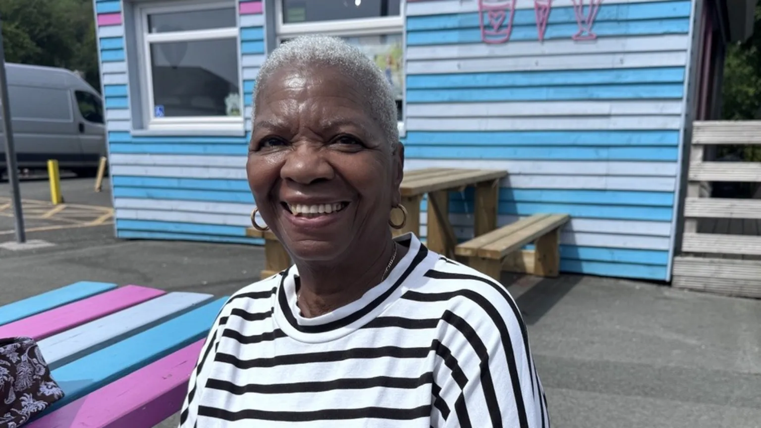 Hortense sat on a bench in front of a blue, white and pink ice cream parlour. She wears a black and white stripey t-shirt and has short blonde hair.