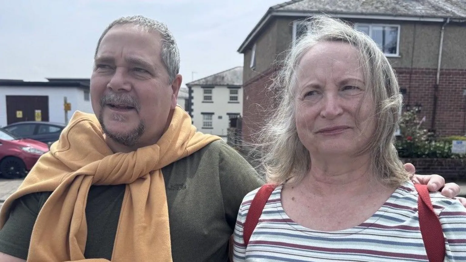 Ian and Andrea looking at the reporter. Ian, on the left, wears a green t-shirt and an orange jumper. On the right, Andrea wears a stripey t-shirt and a red rucksack. 