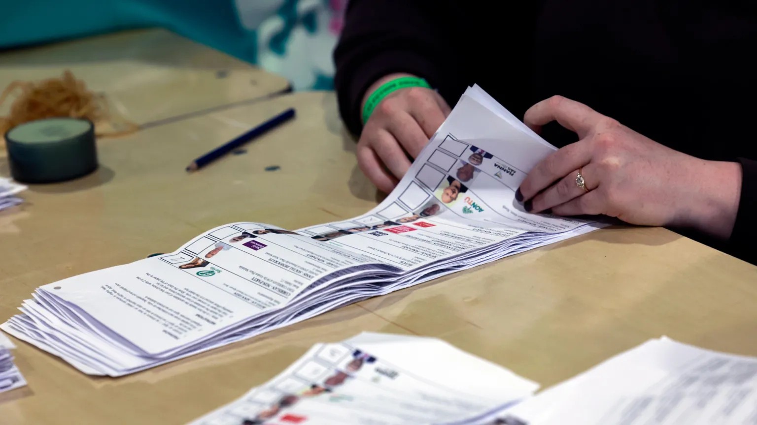 Two hands are visible flicking through a pile of ballot papers on a wooden table.