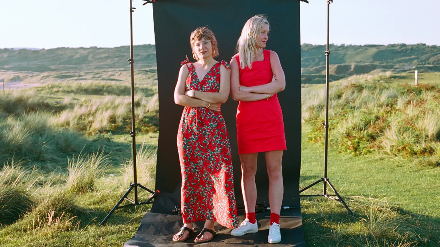 Museum Wales Clémentine Schneidermann and Charlotte James stand side by side in front of a black photography backdrop which is standing in a wild countryside setting of grassy mounds and small hills in the background. Clémentine is left of the picture wearing a long sleeveless orange dress with a grey and white pattern on it, leather thong sandals and a bangle. She has dyed neck-length red hair and is smiling at the camera. Charlotte wears a bright orange mid-thigh fitted dress with white trainers and orange socks. She has dyed peroxide blonde long hair and looks off to the right of the picture.